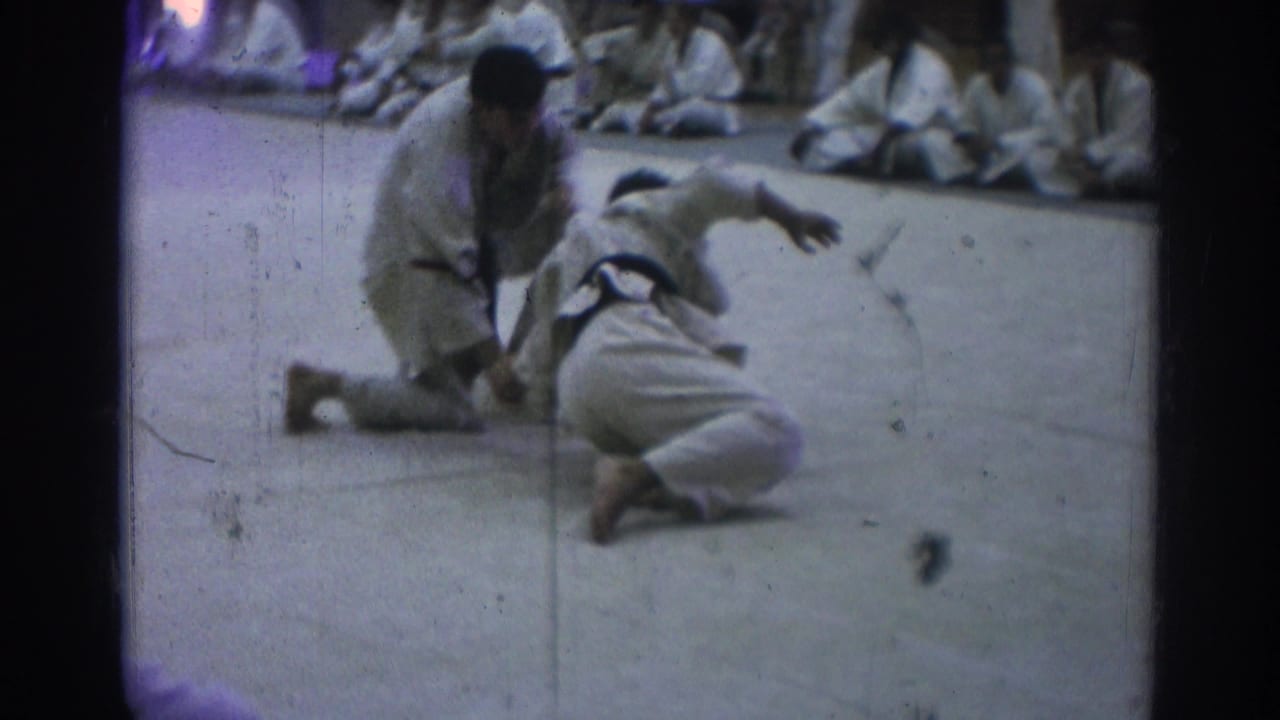 Two martial artists in white gi grapple on a mat while spectators sit behind them.