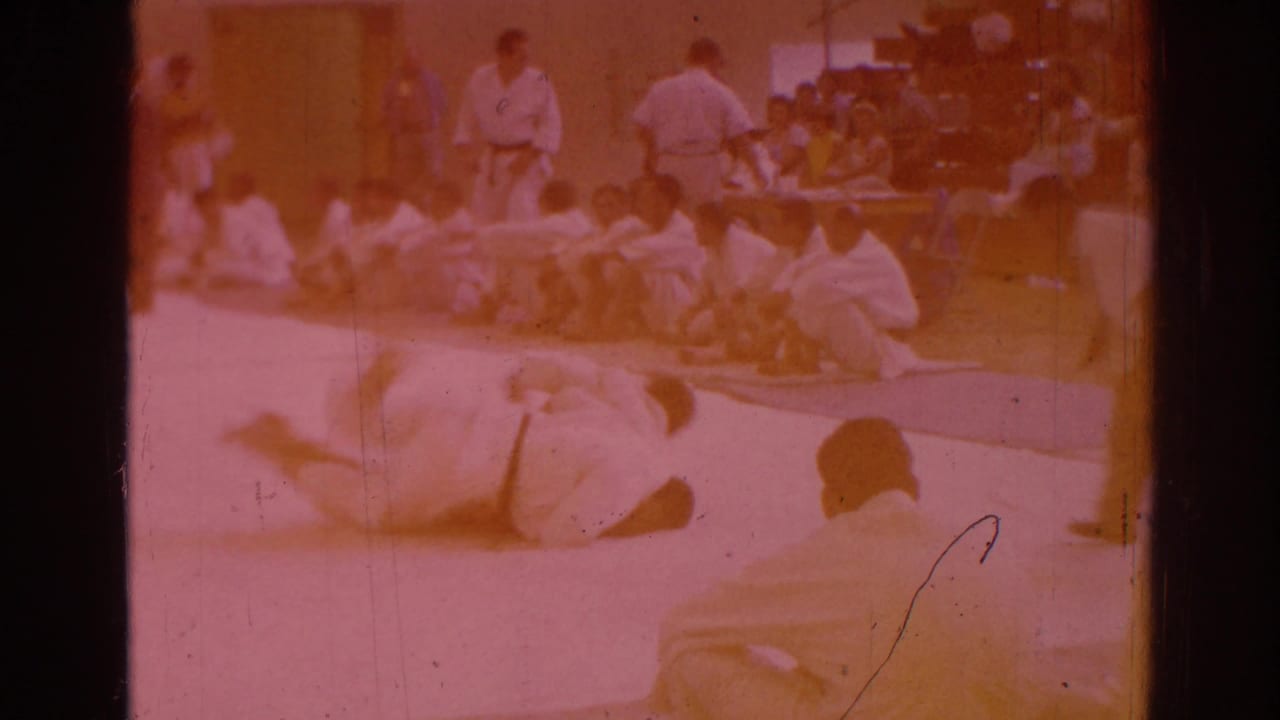 Temple City 1966 competition frame with one martial artist down on the mat and another nearby as seated viewers watch.