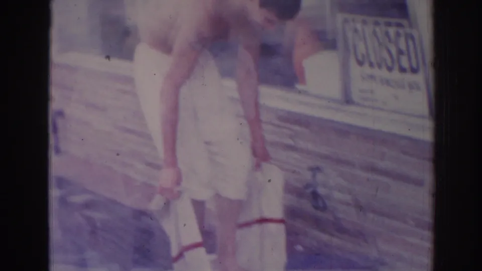The man bends lower beside the brick storefront wall, apparently washing or drying his feet near the base of the window.