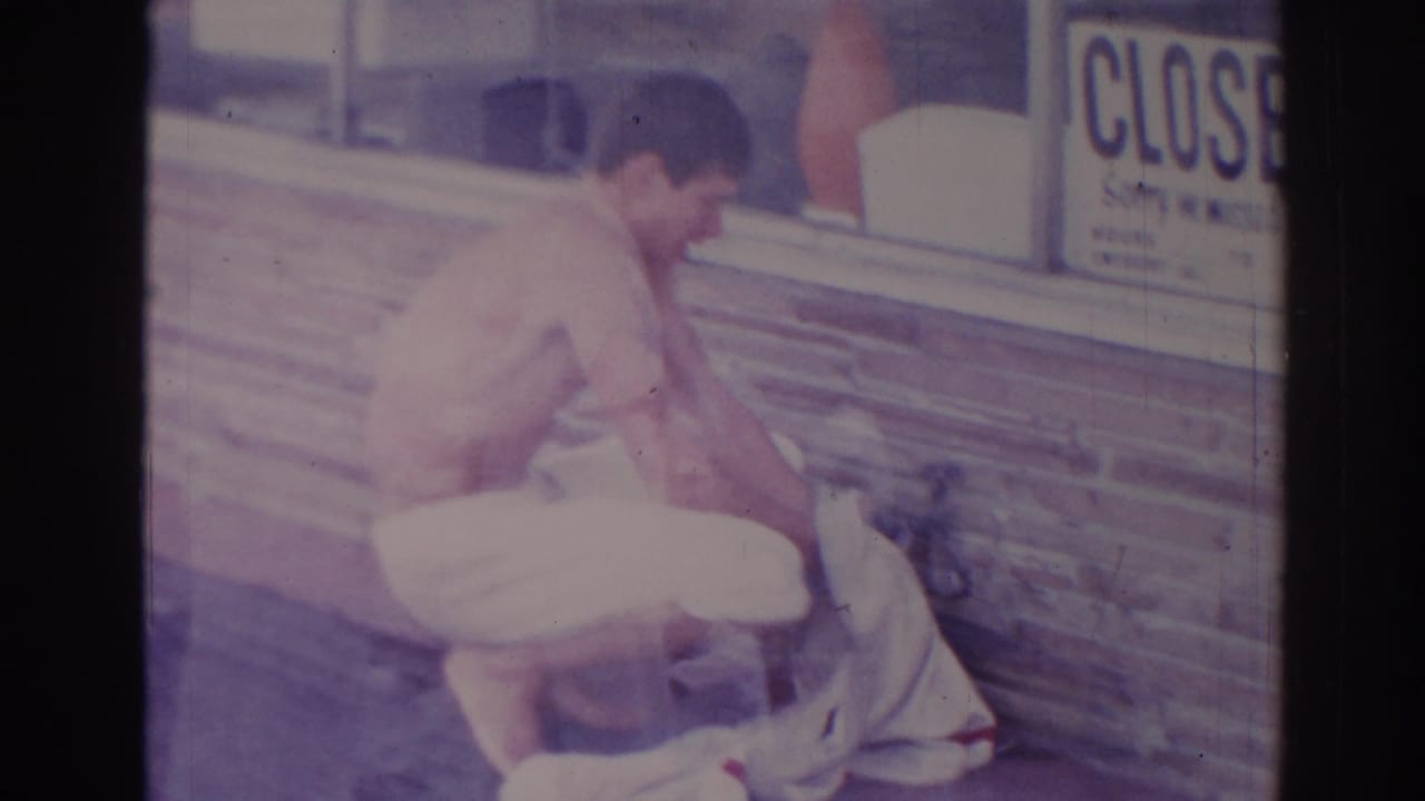 Shirtless man crouches on a sidewalk beside fabric near a storefront window and CLOSED sign.