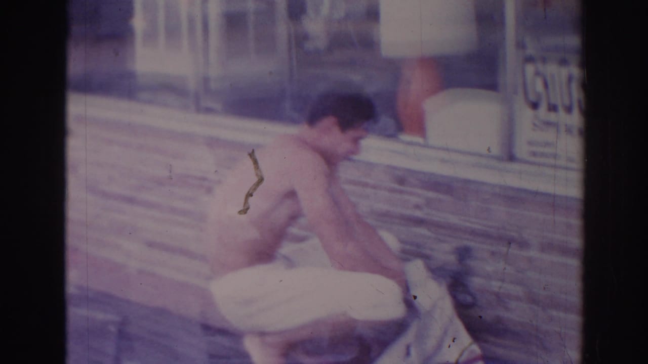 Shirtless man bends over fabric on a sidewalk near a brick storefront and window sign.