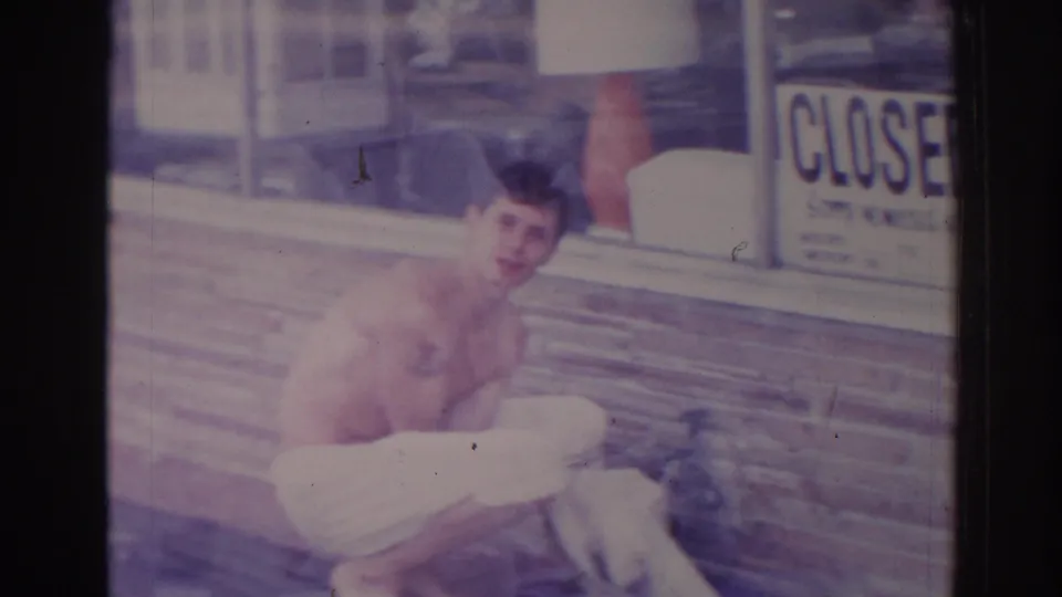 The shirtless man turns toward the camera while crouched near cloth on the sidewalk, with a brick storefront and CLOSED sign behind him.