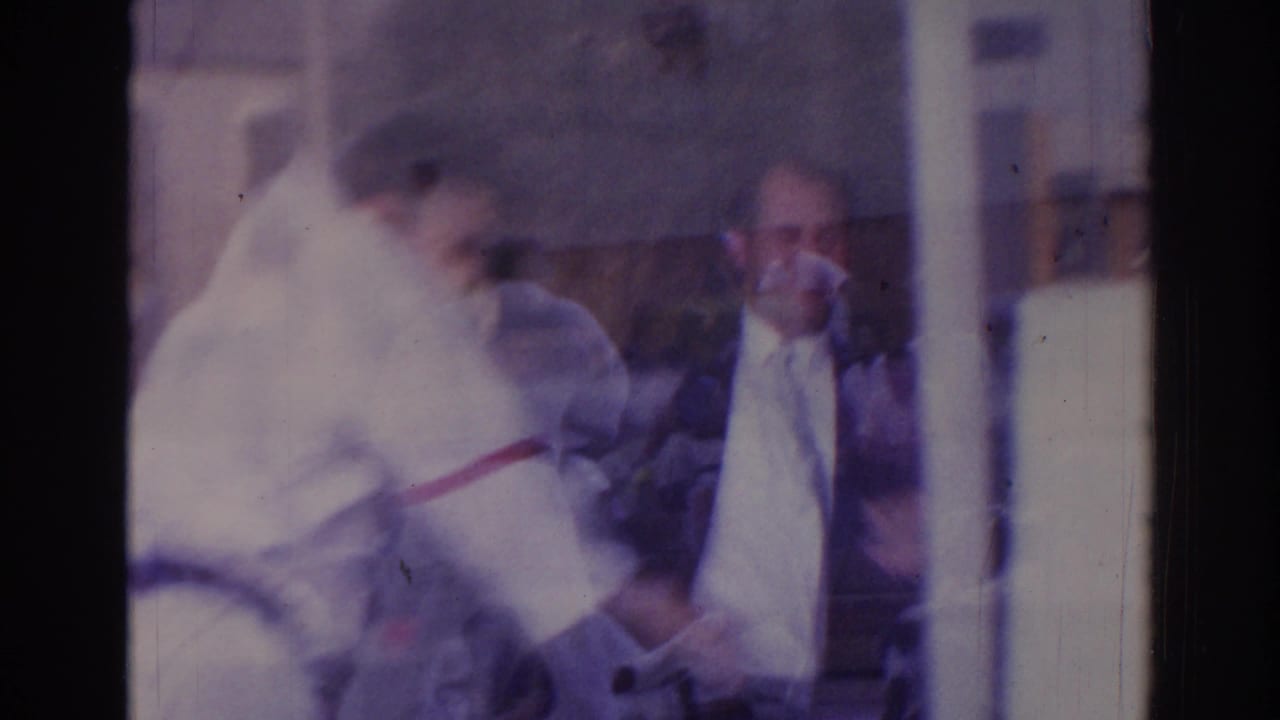 1966 Temple City home-movie frame showing a martial arts practitioner in a white gi and black belt near other people, partly obscured by reflection.