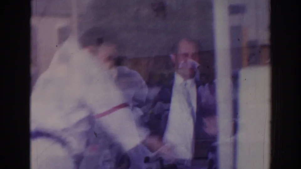 1966 Temple City home-movie frame showing a martial arts practitioner in a white gi and black belt near other people, partly obscured by reflection.