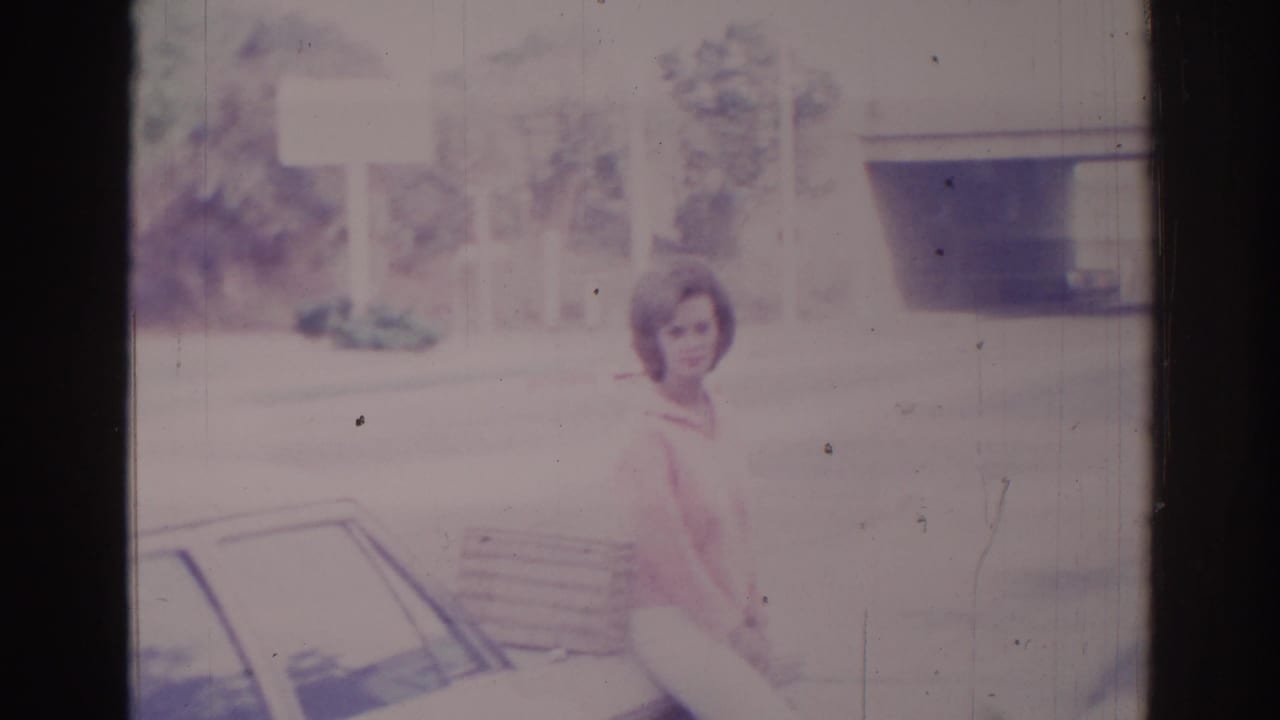 Woman in a pink top stands near a car with a purse in daylight.