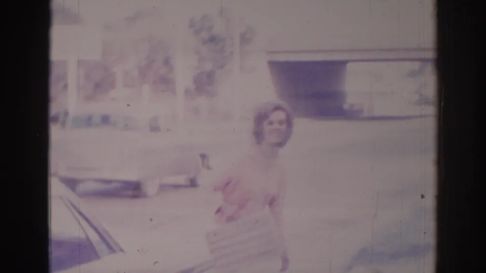 The woman stands beside the car with a woven purse, with a parked vehicle and low building visible behind her.
