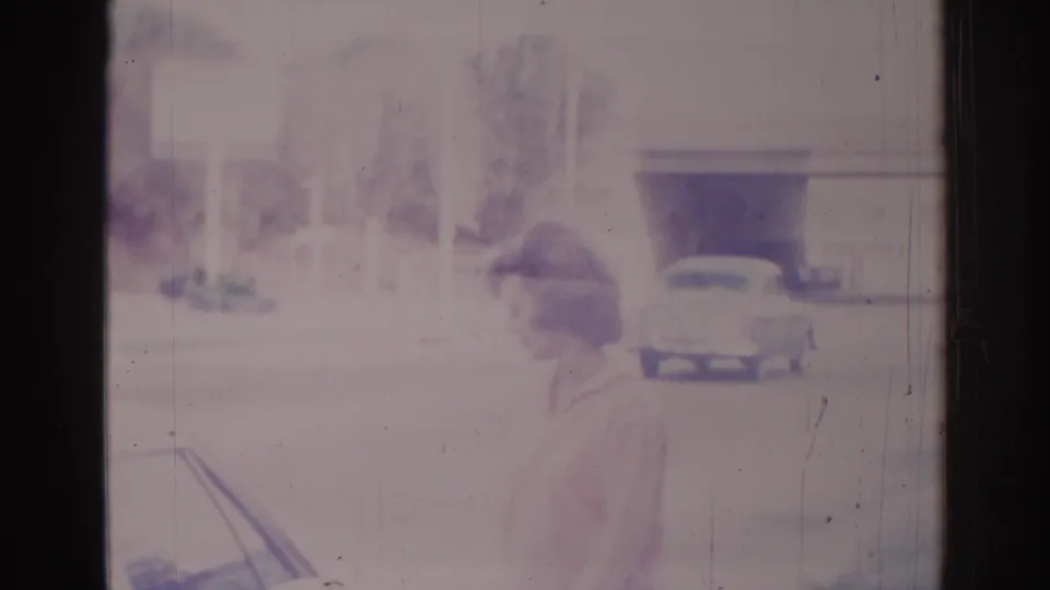 A profile view shows the woman beside the car, with a low suburban building and another parked vehicle in the background.
