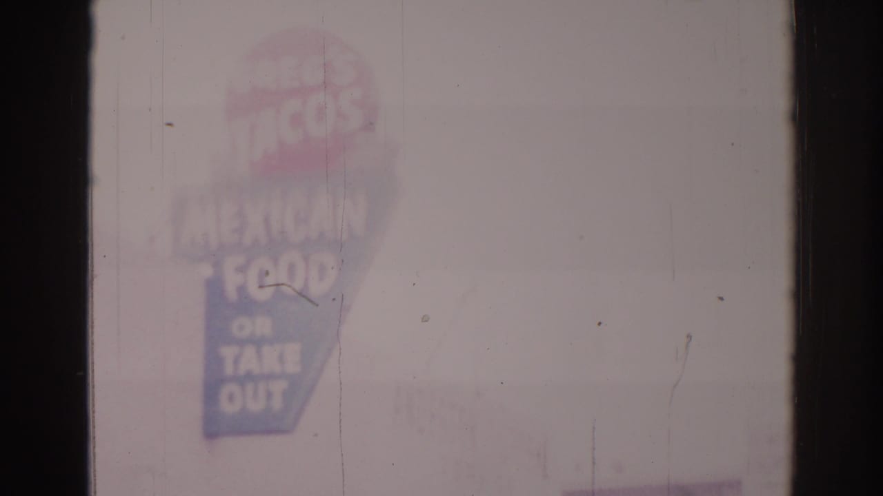 Tall Mexican food takeout sign seen through haze beside a bright street facade.