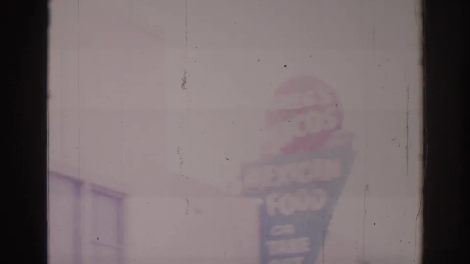Temple City 1966 home-movie still showing the Mexican food takeout sign softened by exposure haze, with a nearby building edge and scratched film surface.