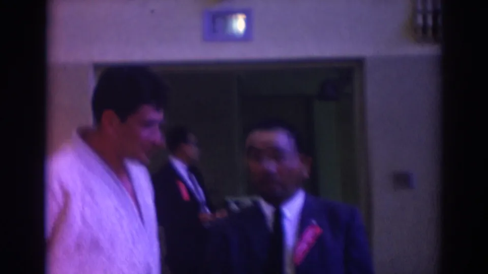 Inside a Temple City building, two adult men appear to converse, one in a white karate gi and one in a dark suit with a red pin.