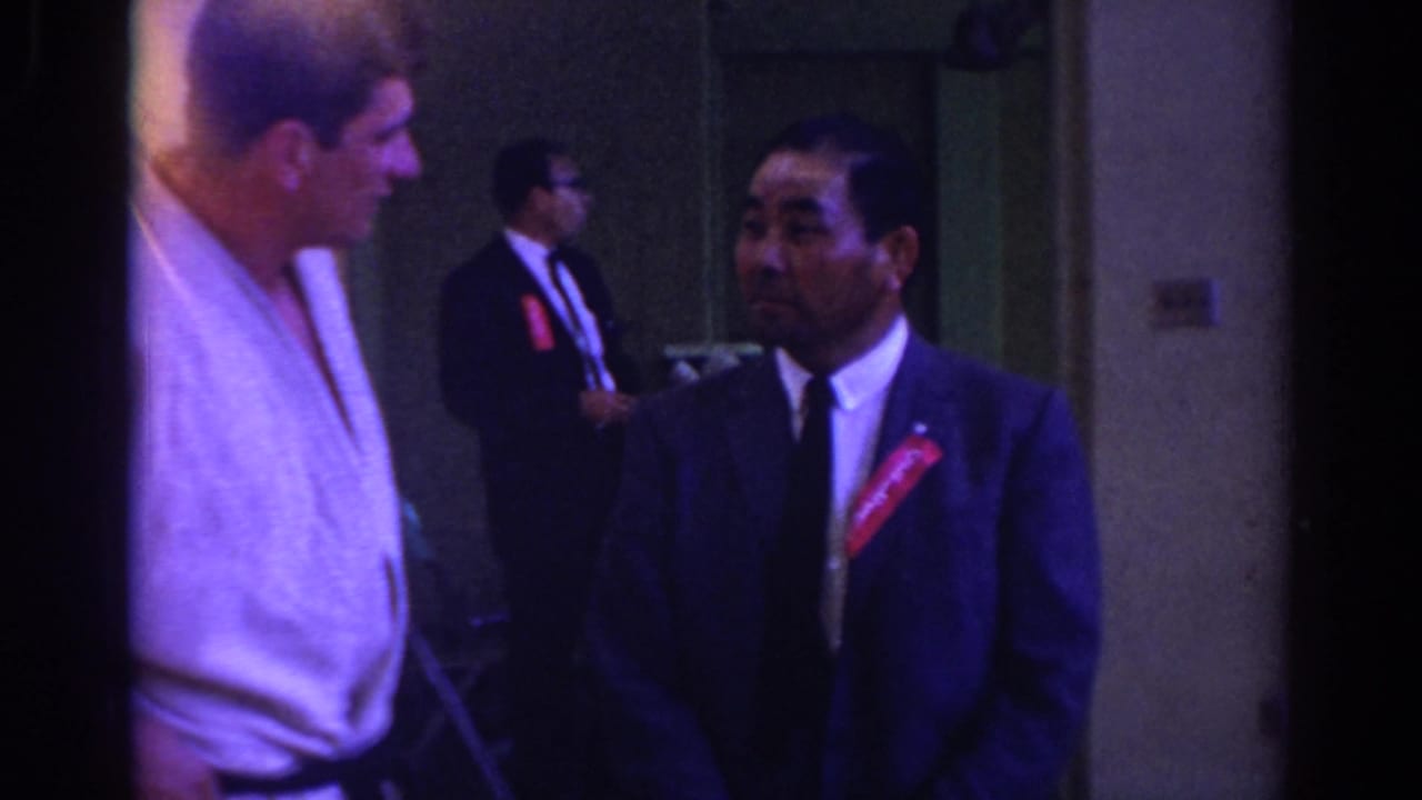 Two men in a corridor, one in a white judogi and one in a dark suit with a red ribbon.