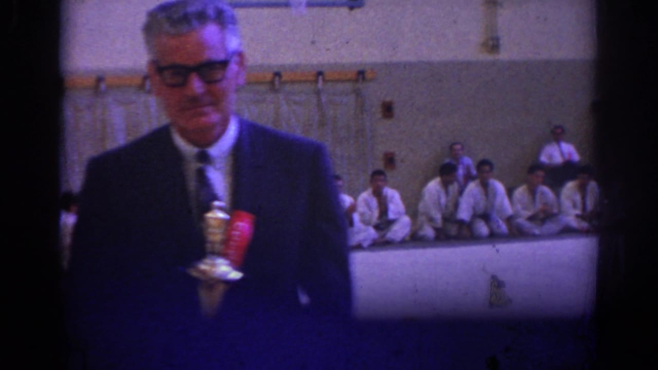 Man in a dark suit and glasses holds a trophy in front of seated students in white uniforms.