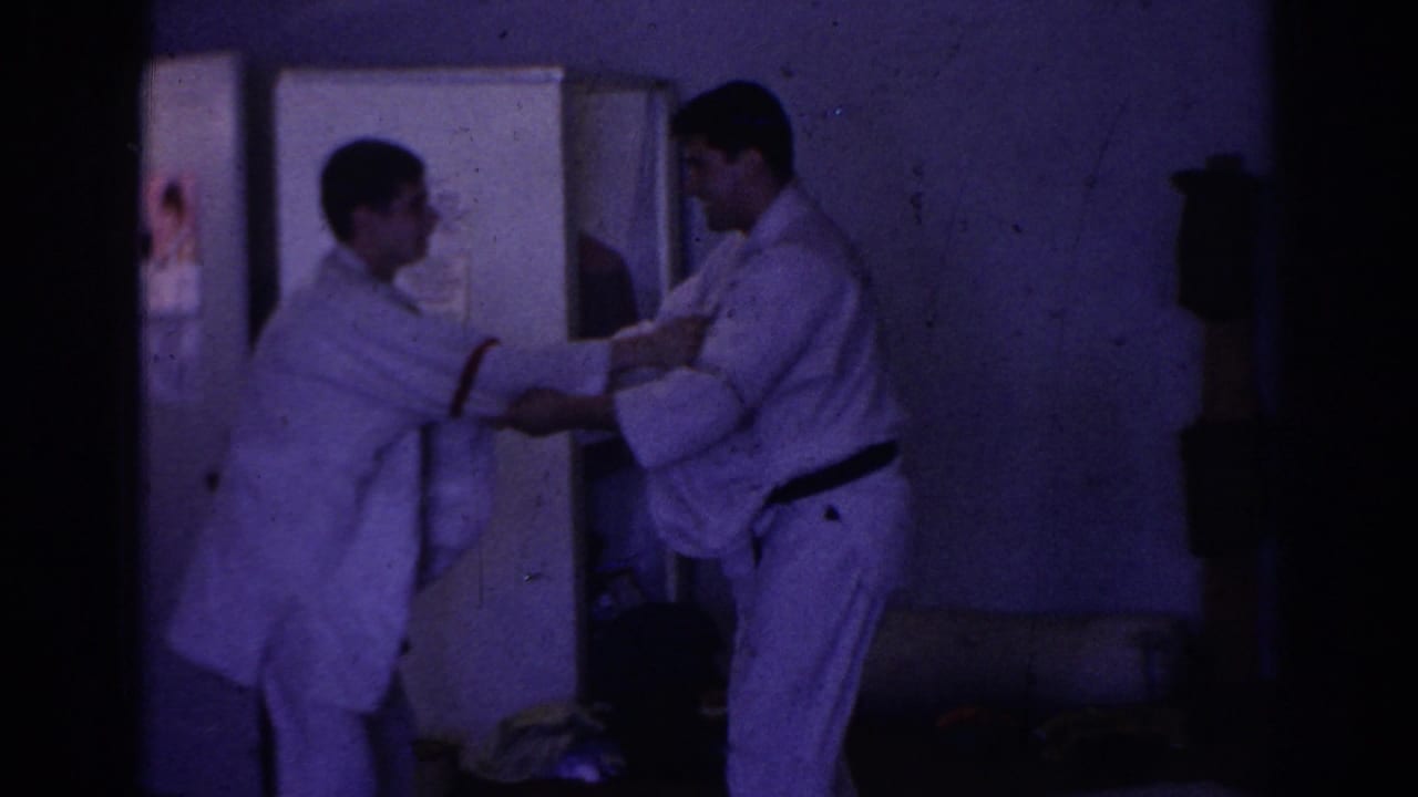 Two men in white gi practice a grip technique in a dim room.