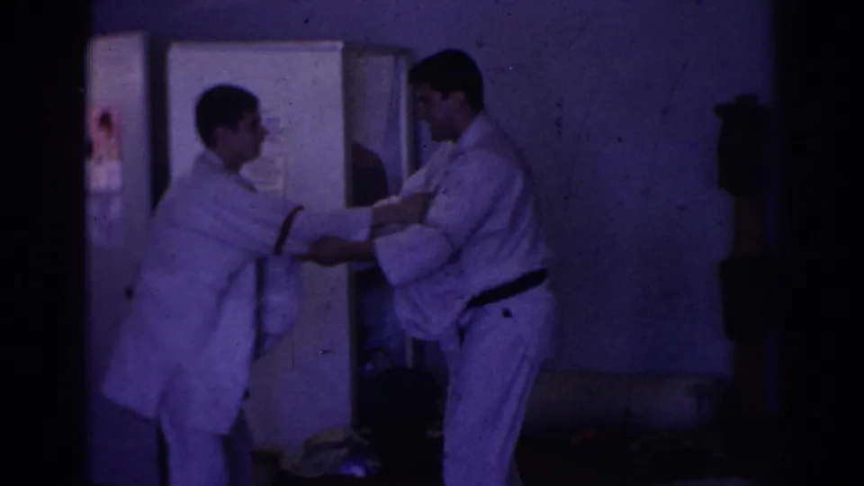 Two men in white gi, one with a black belt, practice a close grip technique inside a dim Temple City interior in 1966 home-movie footage.