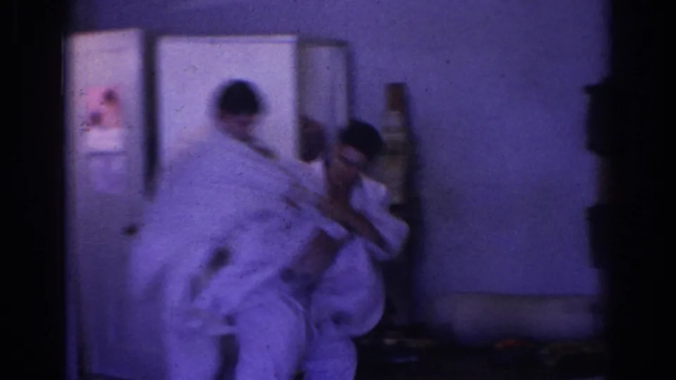Motion-blurred 1966 home-movie frame shows two men in white gi during a close-quarters martial arts movement inside a private room.