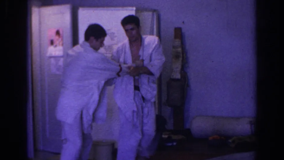 Two men in white gi continue a standing martial arts drill, with a refrigerator, wall post, and padded roll visible in the dim 1966 interior.