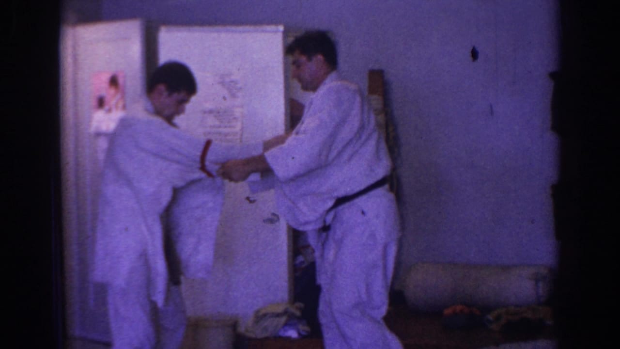 Two adults in white martial arts uniforms practice near a refrigerator in an indoor room.