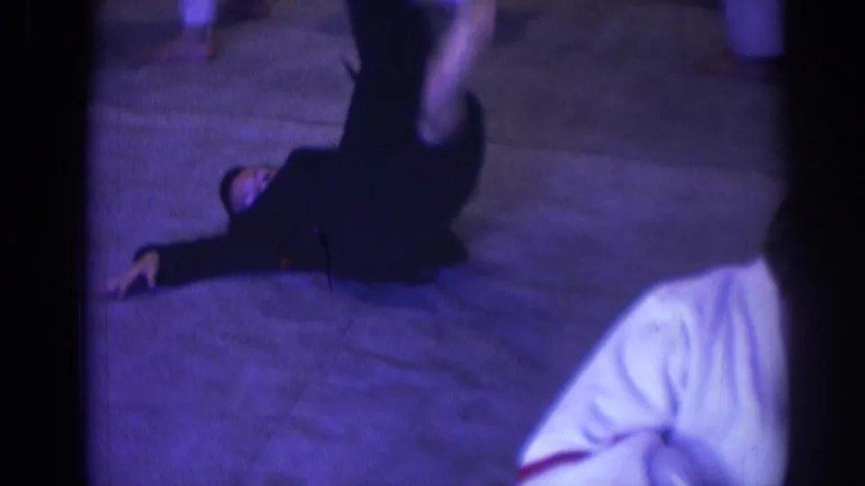 1966 Temple City home-movie frame showing an adult in a dark uniform executing a high kick indoors.
