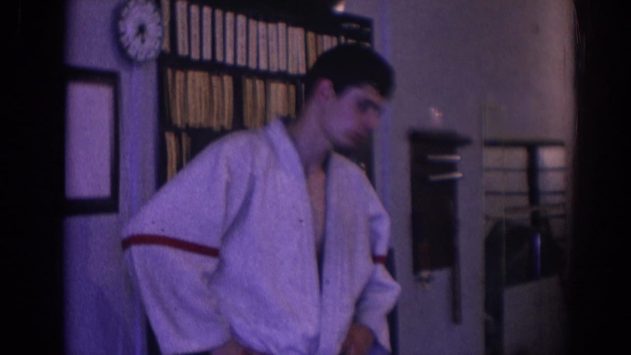 Man in a white gi stands indoors near shelves, a wall clock, and a doorway.