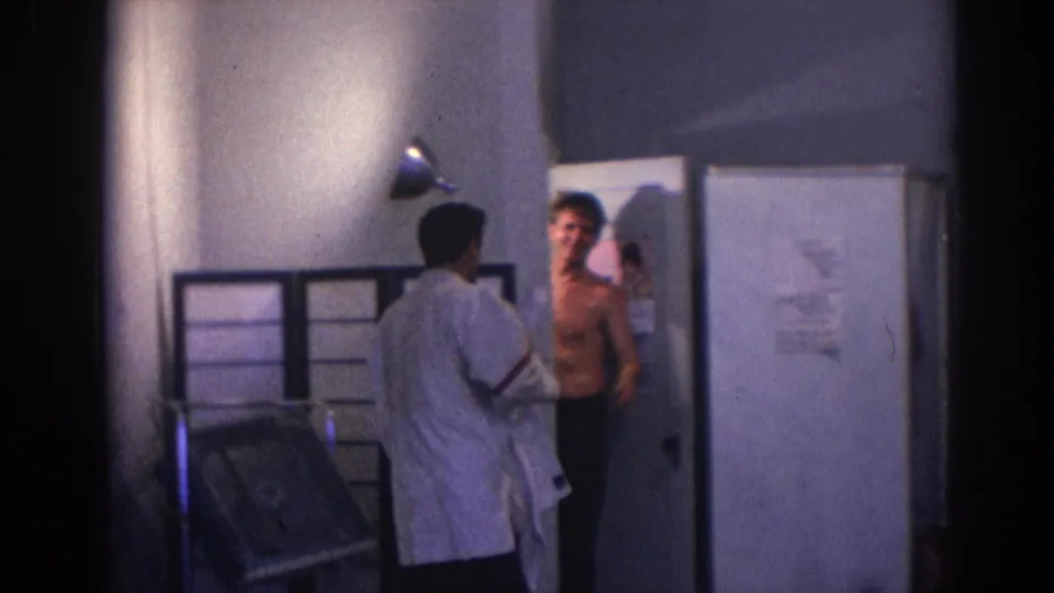 1966 Temple City home-movie frame showing a man in a white gi standing near a shirtless man by a doorway in a private interior.