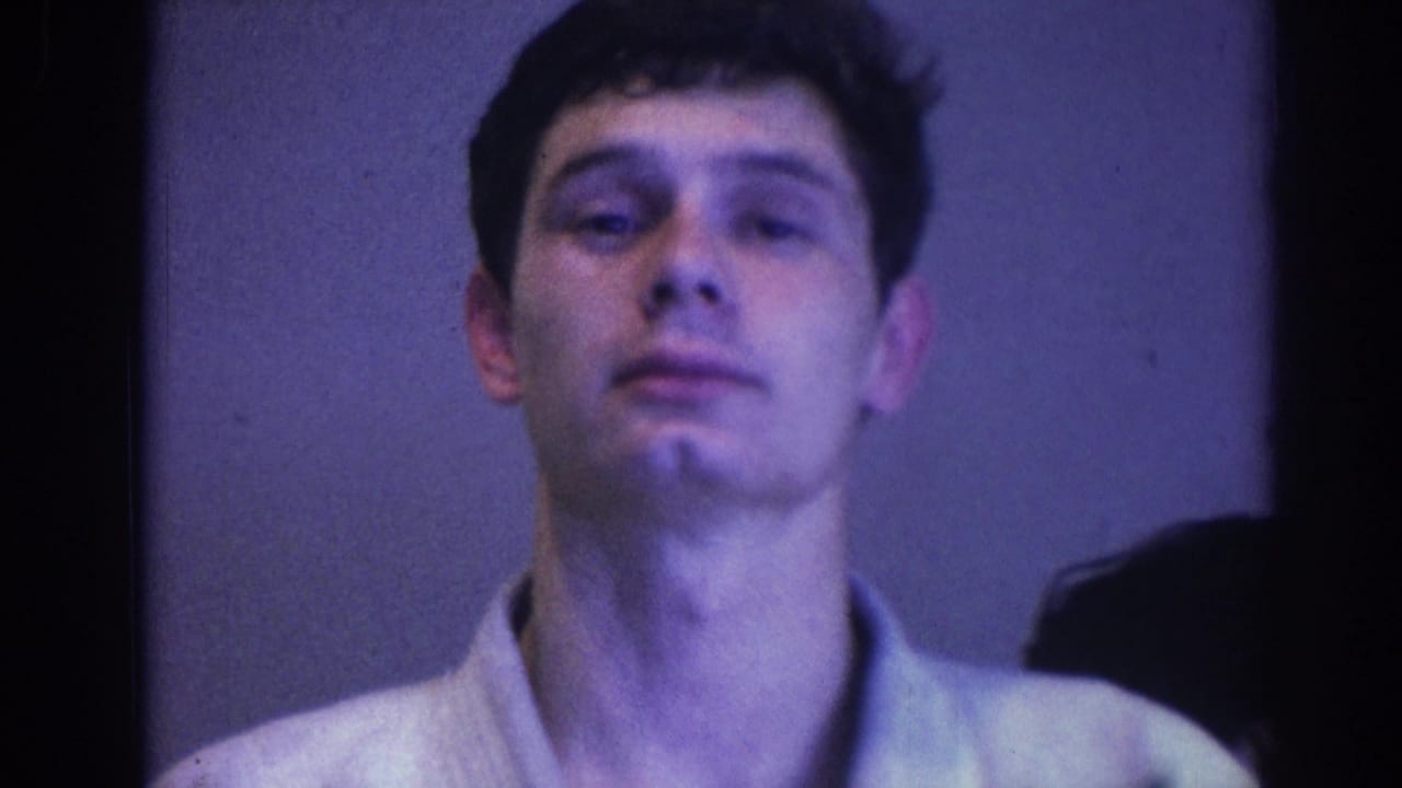 Young man in a light shirt or gi top faces the camera in close-up inside a dim room.