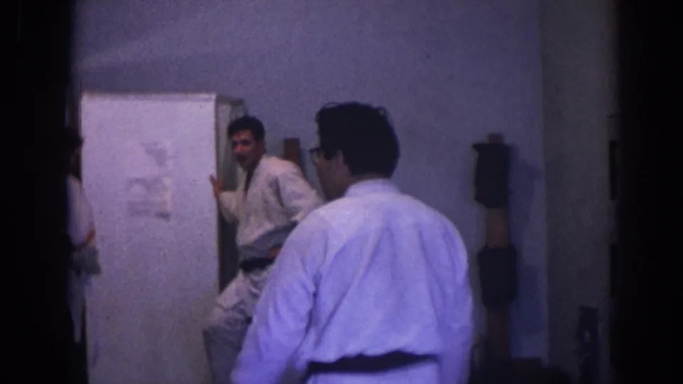 Two practitioners in white gis and black belts appear inside a Temple City dojo-like room, one seen from behind and one near the mirror.