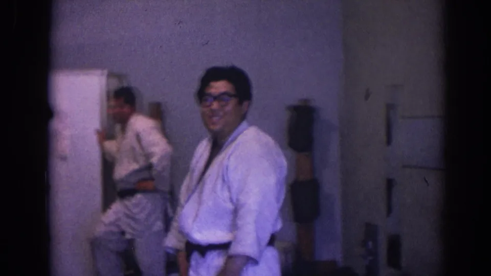A practitioner wearing glasses smiles toward the camera in the dojo-like interior, with another uniformed person reflected or standing near the mirror.
