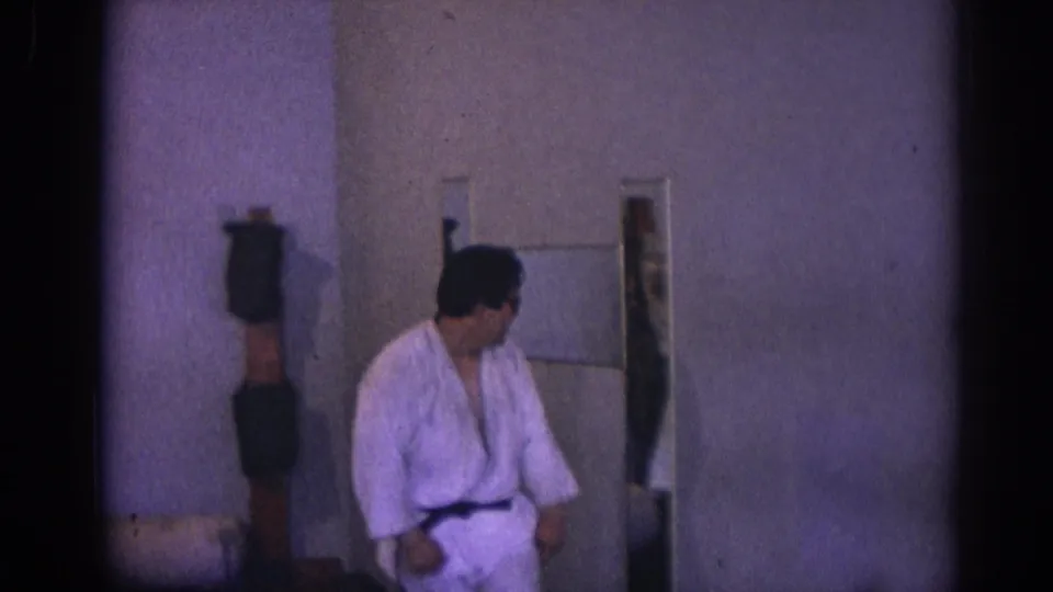 A practitioner in a white gi and black belt stands in the dim Temple City training room beside a wall mirror and padded striking post.