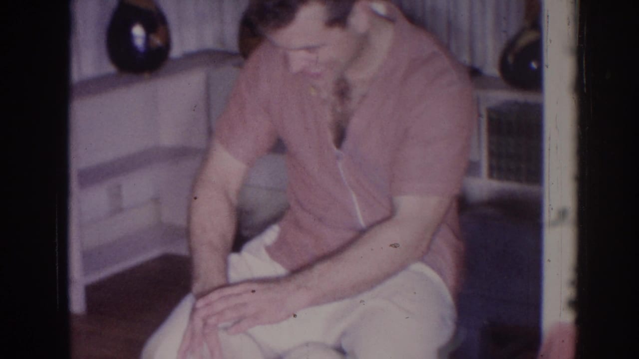 Seated man in a pink shirt holds a small white drum in a living room.