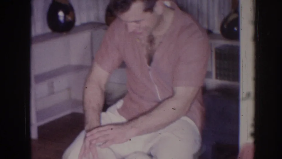 In a 1966 Temple City home interior, a seated man in a pink shirt leans over a small white drum near curtains and a shelf.
