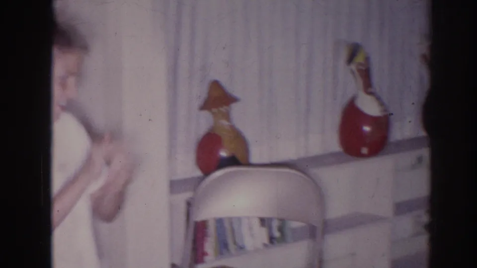 A side view shows the man partly in frame beside curtains, decorative gourds on a shelf, and a folding chair in the home.