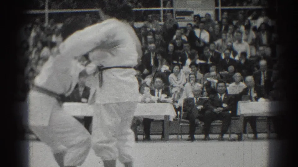 Two martial arts competitors in white uniforms clinch during the West Covina match, with spectators and officials behind them.