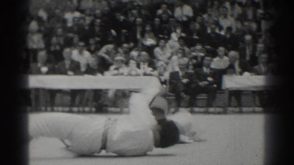 After a takedown in the West Covina match, one competitor lies on the mat with the audience and officials in the background.