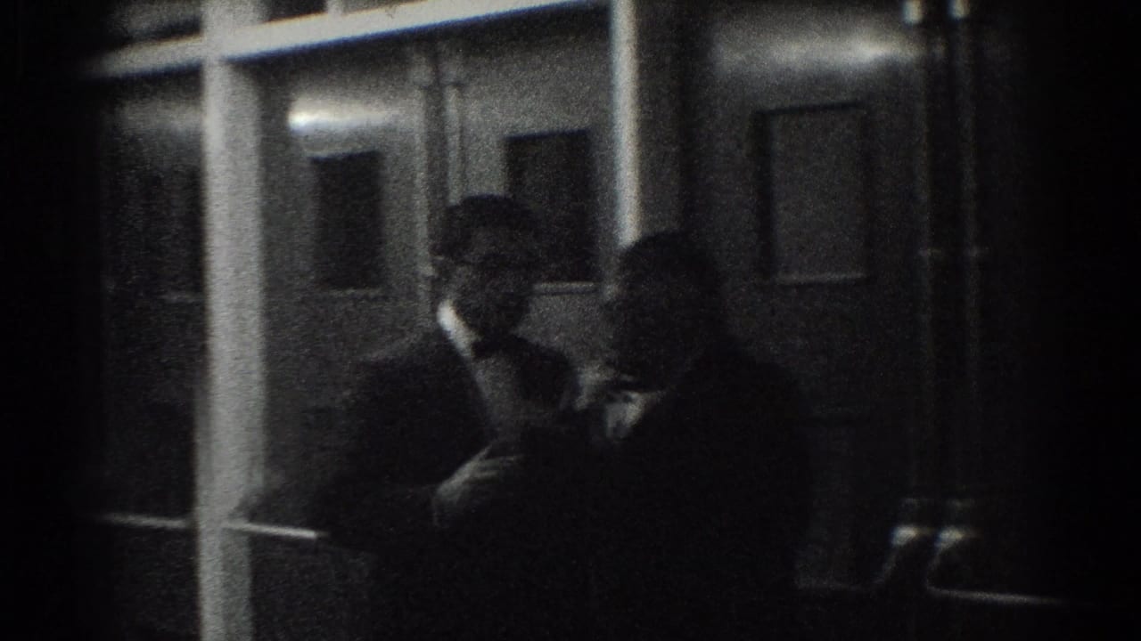 Two men in tuxedos stand in a dim interior near door or window frames.