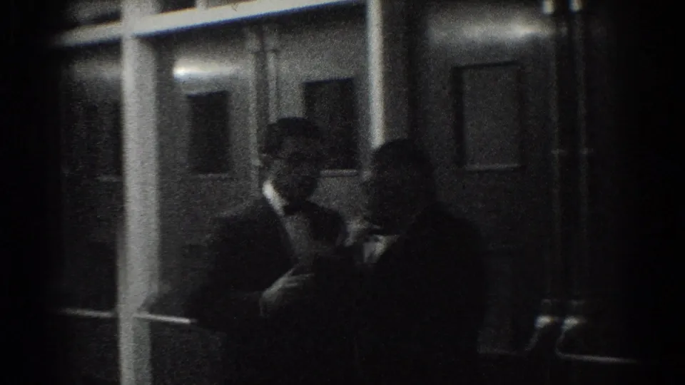 1966 West Covina home-movie frame showing tuxedoed men in a dark indoor setting, one near vertical door or window framing.