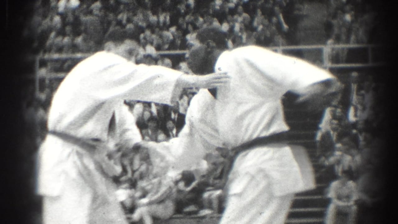 Two judo competitors in white gi grapple on a mat before spectators.