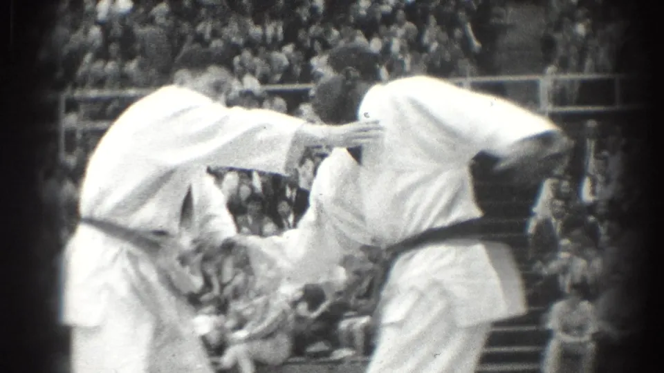 Black-and-white 1966 West Covina frame of judo competitors in white gi and black belts grappling in front of a seated crowd.