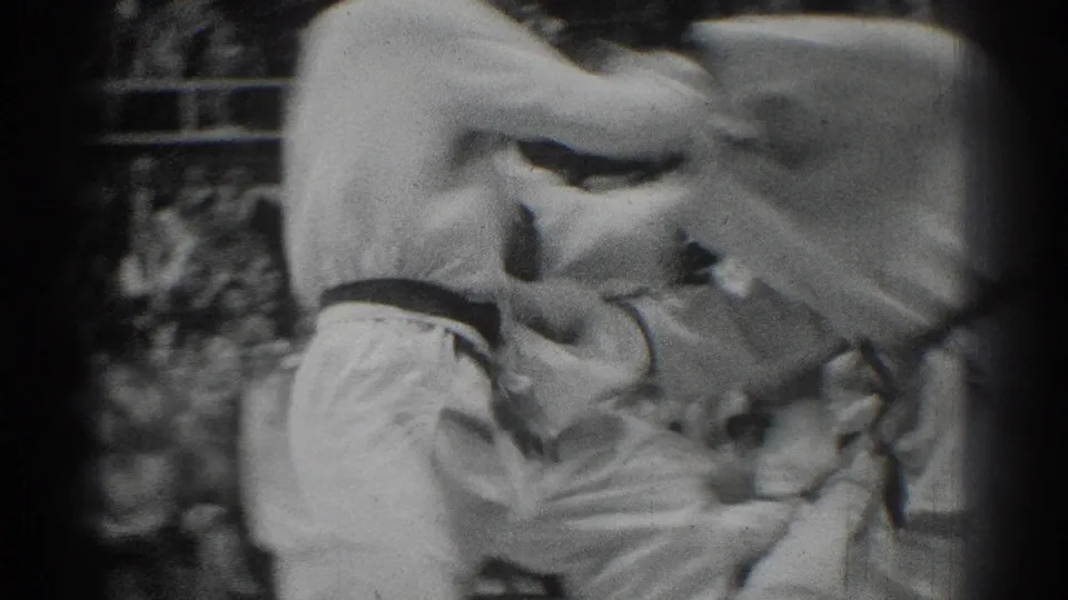 Close black-and-white home-movie view of two judo competitors in white gi during a fast grappling move, with spectators blurred behind them.
