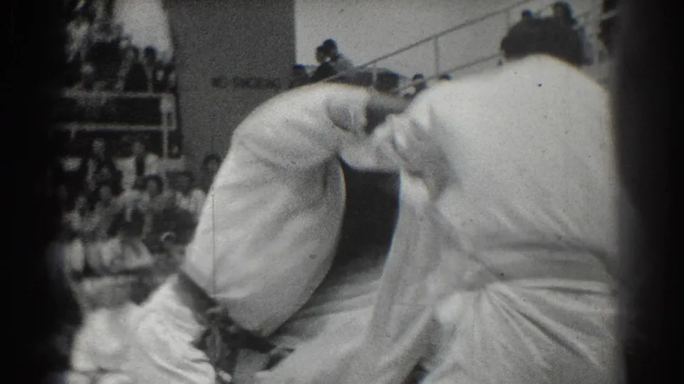 1966 West Covina judo frame showing two competitors in white gi grappling from behind and side angles, with a crowd and railing in the background.