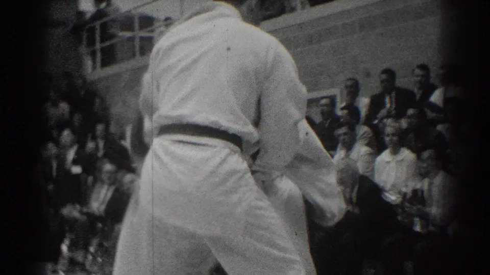 1966 West Covina gymnasium footage shows a karate competitor in a white gi from behind, with spectators seated beyond the mat.