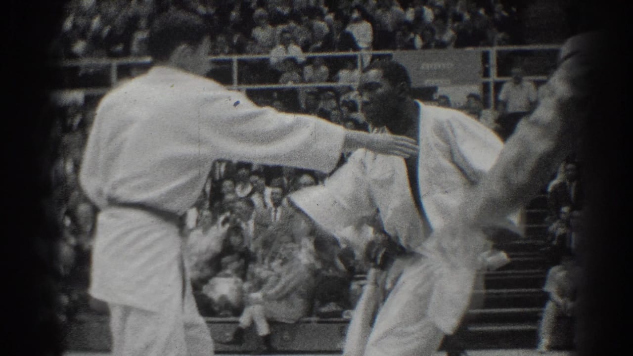 Two men in white karate gi face each other on a mat before a crowd.