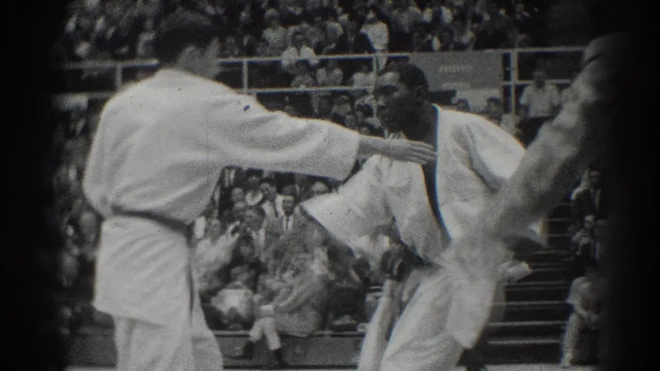 Two karate competitors in white gi face off on a mat in a West Covina gymnasium while spectators watch from bleachers.