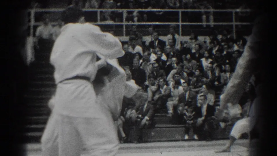The match continues as two competitors in white gi grapple on the gym mat, with a seated audience visible behind them.