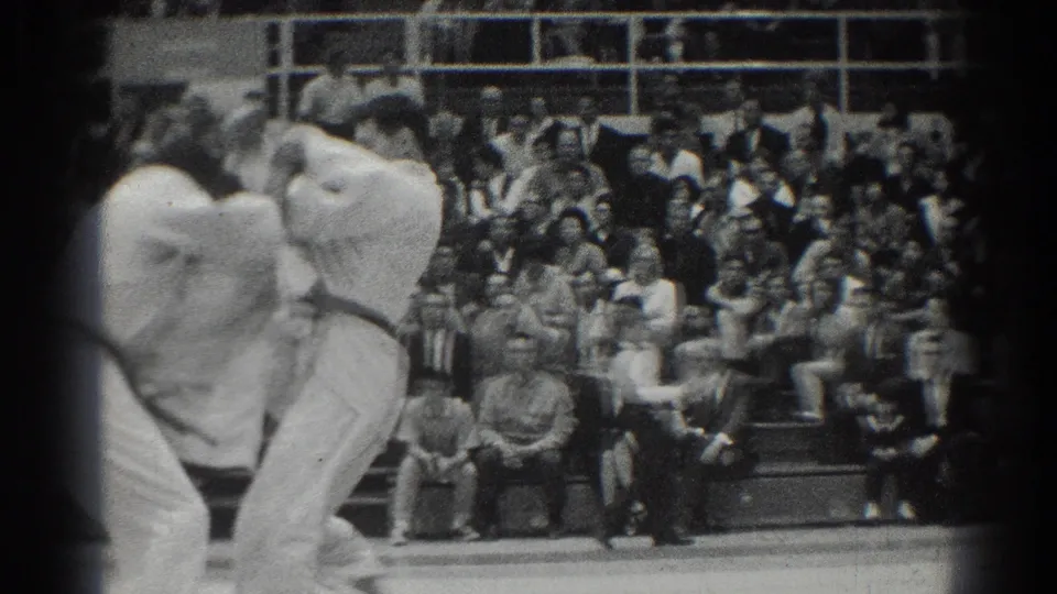 Two karate competitors bend into a grappling exchange on the mat as spectators sit along the gym bleachers in the background.