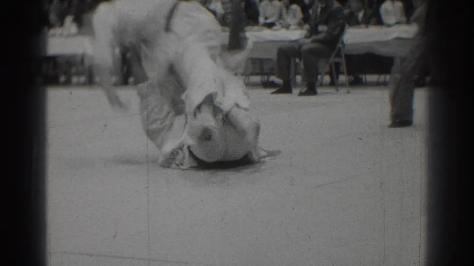 1966 West Covina judo match footage showing two athletes in white gi grappling on the mat before seated officials and spectators.