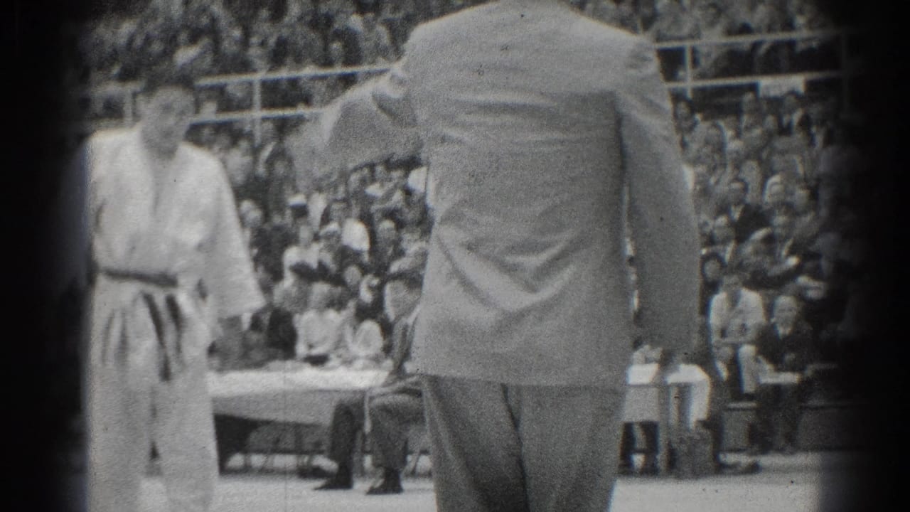A referee stands with his back to the camera near a judo athlete and a crowded gym audience.