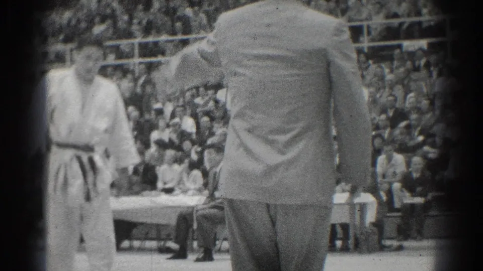 Referee seen from behind during the 1966 West Covina judo championship, with a competitor, officials, and crowd in the gymnasium.