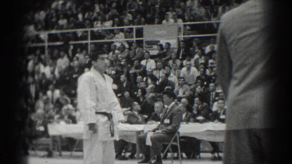 West Covina judo competitor in white gi stands on the mat during the 1966 event, with referee, officials, and audience behind him.