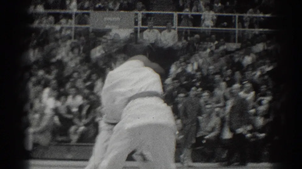 Close spectator-view frame from the 1966 West Covina judo match showing athletes in motion during a throw before a crowded gym.