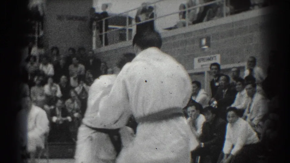 Two martial arts competitors in white gi face off during the West Covina event, with spectators seated and standing in the background.